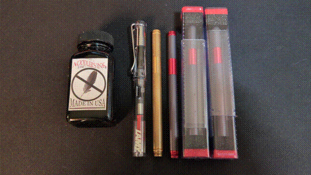 From left to right: A jar of black X-Feather ink, from Noodler's Ink, a transparent plastic LAMY Safari Vista pen with a converter cartridge with a red dial visible, a solid brass TiLiner fountain pen, a dark gray aluminum TiLiner with a red grip, cap tip, and post, and two more of the same still in their plastic boxes.